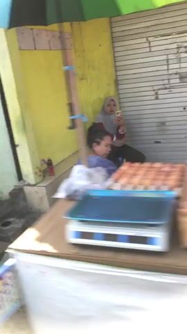 The video shows an outdoor market scene with two people. In the background, a woman wearing a hijab is seated and looking at her phone. In the foreground, a person is seated and appears to be attending to something or resting. In front of them is a digital scale and trays of eggs are visible on a table. The environment seems to be a market stall with a yellow wall and a corrugated metal shutter. The lighting suggests it is daytime.