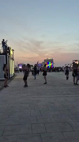 This is a video of an outdoor event, possibly a festival or concert, during dusk. The scene is characterized by a clear, pale orange sky with scattered clouds. In the foreground, a paved walkway leads into a dirt and gravel area where people are gathered. To the left, a modern, black and white modular structure with lights and signage that reads "PLOOOM" is visible, with people on top and in front of it. Further into the scene, several large stages or scaffolding structures are erected, adorned with bright red and blue lights, suggesting a musical performance is either ongoing or about to begin. People are milling around between these stages and structures, some walking in small groups, others standing and observing. The overall atmosphere appears relaxed and social.
