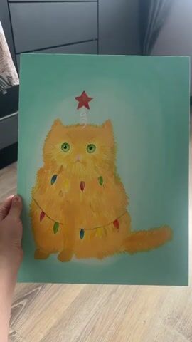 A human hand holds up a vertical rectangular painting depicting an orange fluffy cat adorned as a Christmas tree with colorful lights and a red star on its head against a light teal background. The painting is moved to the left, revealing a real, plump orange cat with green eyes sitting on a light wooden floor. The cat briefly licks its nose and then sits calmly, looking around. In the background, dark grey cabinets are visible, along with a small white bowl featuring fish patterns on the floor.