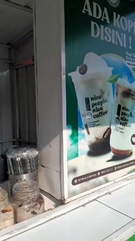 A street-side coffee stand is shown. A banner displays an advertisement for "nice people. nice coffee." Inside the stand, a young man in a gray t-shirt stands behind a counter. Several jars containing ingredients like white powder and brown toppings are visible on the counter. To the left, a container of plastic straws is present. A woman wearing a blue shirt and headscarf is seen working behind the counter, possibly preparing drinks. She is next to an orange machine. In the background, large water jugs are visible. The overall environment appears to be an outdoor food or drink stall.