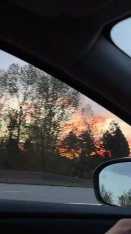 The video captures a view from inside a moving car, looking out the side window at a vibrant sunset in Delta, Canada. The sky is filled with striking orange, pink, and red hues, indicating the early evening or late afternoon.

In the initial frames, two distinct plumes of white smoke or steam rise vertically into the colorful sky from industrial structures visible in the distance, partially obscured by trees. As the car continues to move, more trees and some roadside buildings become visible. Other vehicles can be seen on the road, with one white car briefly prominent. The motion of the car blurs the foreground elements, particularly the trees and passing scenery, emphasizing the sense of travel. The overall impression is one of a scenic drive during a picturesque sunset, juxtaposed with elements of an industrial or developed area.