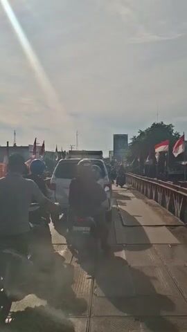 The video shows a view from a bridge, looking forward. The bridge has a metal structure with red and white painted beams. Several Indonesian flags are attached to the railing on the right side of the bridge, blowing in the wind. On the bridge, there is traffic, including cars and motorcycles. In the foreground, a motorcycle with two people is visible. The sky is bright and cloudy, with the sun shining, casting shadows. In the background, there are buildings and trees. The overall impression is a scene of daily life on a bridge in Indonesia, possibly decorated for a national celebration.