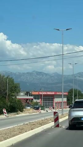 The video shows a view from a vehicle driving on a road. In the foreground, the rear of a silver car is visible. The road has a curb and a barrier. In the background, there is a building with red trim and large garage doors, identified as "TEHNIČKI PREGLED" and "Auto Centar Tomić". Mountains are visible in the distance under a blue sky with scattered clouds. A white car is seen driving in the opposite direction.