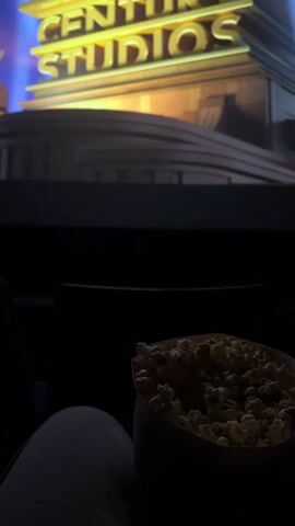 Here is a description of the video:
The video shows a person watching a movie at the theater. The screen shows the 20th Century Studios logo. A bucket of popcorn is on the person's lap. The theater is dark.