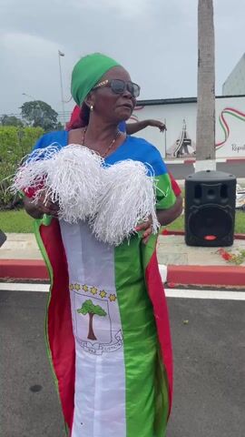 A woman wearing a green headscarf, sunglasses, and a dress adorned with the flag of Equatorial Guinea stands outdoors, holding two white pom-poms. The dress prominently features the country's coat of arms, including the silk-cotton tree, six stars, and the motto "UNIDAD PAZ JUSTICIA" (Unity, Peace, Justice). She smiles and moves rhythmically, possibly dancing, to music emanating from a visible speaker on the right. Another woman, similarly dressed in a colorful outfit resembling the national flag and wearing a red headscarf, stands behind her, also moving. The scene appears to be a public area or street, with paved surfaces, a curb, and some greenery. In the background, a wall displays a design incorporating the flag colors and partially visible text, "Republica de Gu...", likely referring to "Republica de Guinea Ecuatorial." The sky is overcast, indicating a cloudy daytime setting. The overall mood appears celebratory or festive.