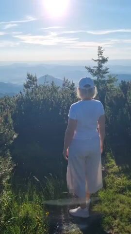 A woman wearing a white t-shirt, white cap, and light-colored pants walks away from the camera on a dirt path through a mountainous landscape. She is walking towards a bright sun in a hazy sky with scattered clouds. On either side of the path are dense, green bushes and evergreen trees. The path appears to be on the side of a mountain, with rolling hills and more distant mountains visible in the background. The lighting suggests it is either sunrise or sunset, with the sun low in the sky and casting a warm glow. The overall mood is peaceful and serene, evoking a sense of solitude and connection with nature. There is no visible text in the media file.