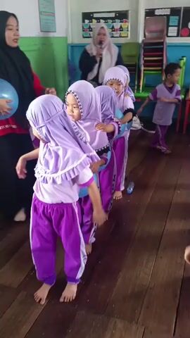 In an indoor classroom setting with wooden floors, a group of young children, dressed in matching purple uniforms and headscarves, are participating in a game. They stand in a line, holding blue balloons between their knees. A woman wearing a black hijab and a red shirt is interacting with the children in the foreground, seemingly guiding or instructing them. Another woman in a light-colored hijab is visible in the background, holding a microphone and speaking. The environment appears to be a kindergarten or preschool, with educational posters and colorful toys on display. The children are focused on holding the balloons as they move or stand in formation.
