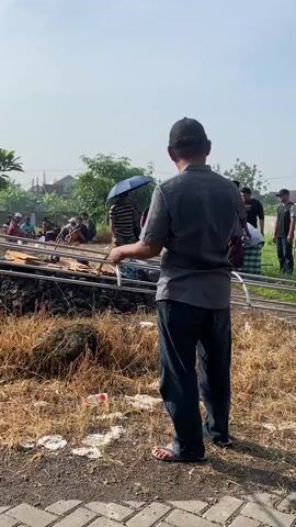 The video shows a group of people gathered around a grave site in what appears to be a rural or semi-rural area. Some individuals are standing near a grave while others are actively digging with shovels.  There is a metal casket frame or bier resting near the grave. A few people are sitting or kneeling around the grave site, and some are holding umbrellas. A man in a cap and casual attire is walking in the foreground, holding a folded cloth, while another man in a green shirt passes by. The environment includes dry, yellowed grass, scattered trees, and a building resembling a mosque or religious structure in the background. Overhead, there are electrical wires.