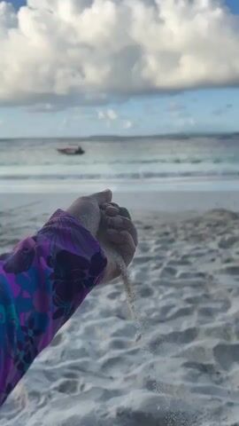 This is a close-up, vertical, first-person perspective video of a hand, wearing a purple and blue floral patterned long-sleeved shirt, holding sand in its cupped palm. The hand is slowly releasing the sand, which falls through the fingers and dissipates in the air. The background shows a beach with white sand and sand dunes, and beyond that, the ocean with small waves and a boat in the distance. The sky is cloudy with patches of blue. The overall lighting suggests it is either early morning or late afternoon.