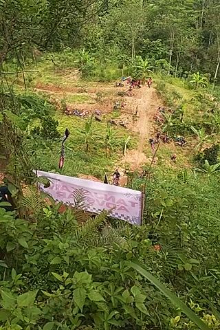 The video captures a steep, muddy dirt path winding through a heavily vegetated, hilly landscape. Lush green trees and various plants cover the slopes, suggesting a tropical environment. The scene is illuminated by daylight, indicating sunny weather.

In the foreground, a white and purple banner with partially visible text, possibly reading "SAKAYON" and other words, hangs among the foliage. Several ropes are strung across the area.

A person wearing a white helmet and dark clothing is seen carefully navigating the challenging, muddy path. Further up the slope, multiple dirt bikes or motorcycles are visible, some parked and others with people around them, suggesting an off-road event or trail ride. Other individuals are also present along the path. The overall activity appears to be an adventurous and physically demanding outdoor event taking place in a natural setting in Kerjo, Indonesia.