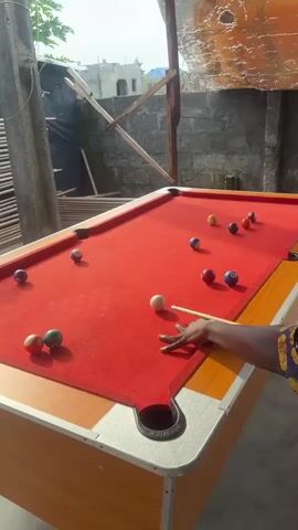 A person's hand is visible, holding a pool cue and poised to strike a white cue ball on a red-felted pool table. Several colored billiard balls are scattered across the table. The scene appears to be outdoors, with a brick wall and some makeshift structures in the background. The lighting suggests it is daytime.