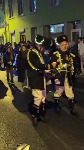 The video shows a festive parade taking place in a town or city at night. The parade includes people dressed in traditional clothing with festive lights, some playing instruments such as accordions and drums. Two men in black military-style jackets with gold trim and lit hats are at the front of the group. The street is lined with people watching the parade, and the buildings on either side are painted in bright colors. The street lights provide additional illumination. The atmosphere seems joyous and celebratory.
