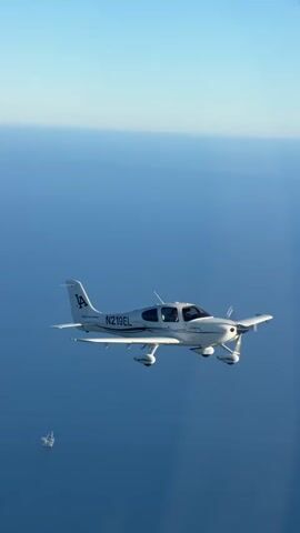 This video is an aerial shot of a white single-engine airplane flying over the ocean. The airplane is positioned in the center of the frame and is seen from the side. It has the registration number N219EL clearly visible on its fuselage. In the background, the vast expanse of the blue ocean stretches out to the horizon, with a sliver of lighter blue sky visible above. To the far left and slightly below the airplane, a small, distant boat or ship is faintly visible on the water. The perspective suggests the video was filmed from another aircraft or a drone.