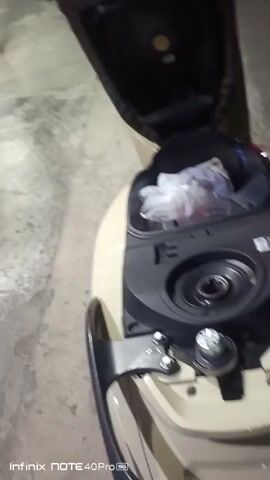The video begins with a close-up of a motorcycle's storage compartment, which is open and contains a few items wrapped in plastic bags. The camera then pans to the left, revealing a person standing near a concrete floor. The person is wearing a t-shirt with a palm tree design, black shorts, and flip-flops. They are holding a small, dark object in their hand, possibly a phone. The scene appears to be indoors, possibly a garage or workshop, with some wooden structures and bamboo poles visible in the background. The lighting is dim. The camera then moves towards a red and white gas pump, branded "Pertalite" and "FULL INJECTION." The gas pump has a digital display showing prices and quantities. The person is now seen walking towards the gas pump, and the camera focuses on the gas pump itself. The digital display shows "RP 12.000" and "L 1.00." The person's legs and feet are visible as they move near the pump. The video ends with a shot of the gas pump, with the person partially visible in the background. The environment suggests a service station or a similar facility.