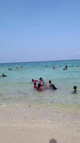 The video shows a beach on a clear, sunny day. Several children are playing in the shallow, turquoise water near the shore. Other people, including adults and children, are swimming further out. The beach is sandy and the water is clear enough to see the sandy bottom. The overall atmosphere seems relaxed and joyful.