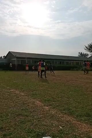 A group of people, likely students, are gathered on a grassy field in front of a long, single-story building. The building has a white facade with a red stripe and many windows. The sky is partly cloudy with bright sunlight filtering through. The individuals are dressed in athletic attire, with some wearing orange shirts and yellow shorts, and others in green vests. They appear to be engaged in some form of physical activity or training, with one person in the foreground performing stretches. The field is uneven and dry in some areas, indicating it might be a sports field or a general outdoor area.
