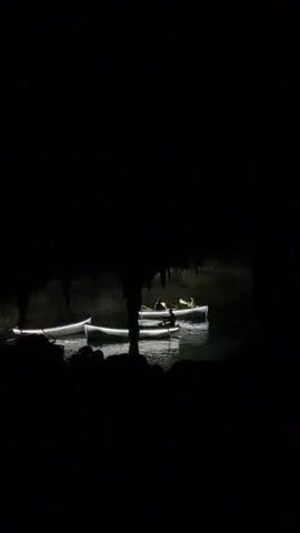 The video shows three illuminated boats floating on a dark body of water. The boats are white with a strip of LED lights outlining their edges. Each boat has two occupants, their figures silhouetted against the faint light. The boats are moving slowly, with one occupant in each boat rowing. The scene is partially obscured by what appears to be the entrance of a dark cave, with stalactites hanging down from the ceiling. The overall environment is dimly lit, with the only significant light source being the LED strips on the boats.