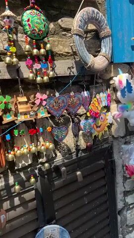 The video pans across a small shop filled with souvenirs and novelty items. On the left, a life preserver hangs on a stone wall, adorned with a welcome sign. Below it are heart-shaped decorations and a colorful sun image with the word "Welcome" written in rainbow letters. Further down, wind chimes hang, and a closed wooden shutter is visible. To the right, a variety of plush toys, including unicorns, a mermaid doll, and a Mickey Mouse plush, are displayed. Next to these are plastic horns and other small toys. The camera then moves to show the interior of the shop, revealing a woman in a white dress browsing items. The shelves are packed with various trinkets, including small figurines, jewelry, keychains, and decorative plates. A rack of colorful items, possibly scarves or textiles, is also present. The shop appears to be an outdoor market stall or a small, independently owned store, with a rustic, stone and wooden facade. The lighting suggests it is daytime, with bright sunlight casting shadows.