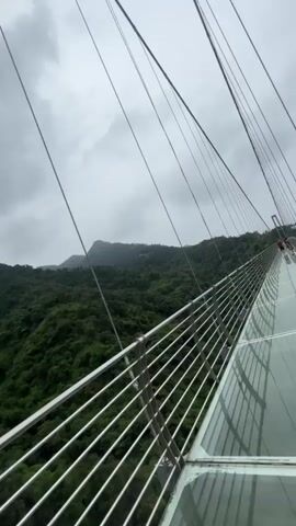 A video captures a person walking on a glass-bottomed suspension bridge, looking out at a lush, green mountain landscape under a cloudy sky. The bridge has metal railings and is supported by cables. In the distance, a large pylon with Chinese characters is visible, as well as some construction scaffolding on a hillside.