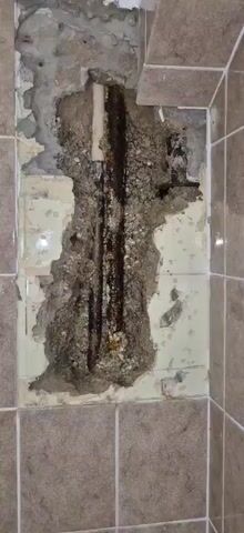 The video shows a close-up view of exposed plumbing pipes within a wall cavity. One pipe is white and appears to be PVC, while the other is metallic and heavily corroded with rust and mineral deposits. The surrounding wall material is rough concrete or plaster, with areas of damage and missing sections that reveal the pipes. There are also signs of moisture and leakage, indicated by dark staining and visible water droplets on the pipes and surrounding material. The lighting is somewhat dim, with the focus primarily on the pipes and the damaged wall. The overall impression is one of decay and disrepair related to plumbing infrastructure.