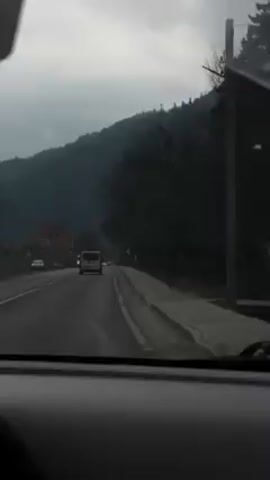 A car drives on a road on a cloudy day. There are other vehicles in front. On the left side of the road is a red brick building with the number 416 on it. Trees line the right side of the road, and a mountain is visible in the background. The windshield wipers are active.