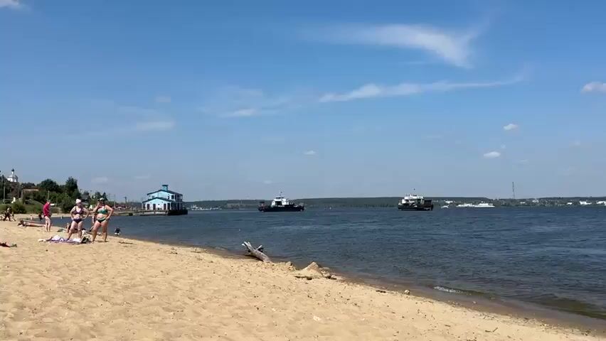 The video shows a wide, calm body of water under a bright blue sky with scattered clouds. In the foreground, a sandy beach with some driftwood is visible. The middle ground features a large river or lake with three vessels. On the far left is a stationary, light blue and dark blue building on a barge. In the center and to the right are two ferries. The ferry in the center is dark with a white and blue superstructure and appears to be moving. The ferry on the right is also dark with a white and blue superstructure and seems to be stationary or moving slowly away from the camera. The ferries have ramps at the front and back. The background consists of a dense, dark green forest line along the distant shore, with some industrial structures and chimneys visible behind the trees. Red buoys are scattered in the water. The overall atmosphere is peaceful and depicts a sunny day on the water.