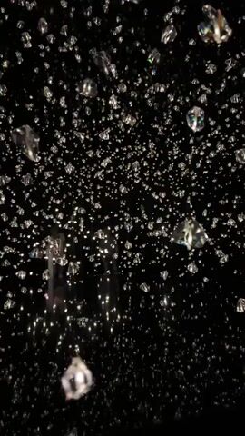 The video shows a dark environment filled with numerous small, faceted objects that appear to be diamonds. These objects are suspended in the air and are catching and reflecting light, creating a sparkling effect. In the background, there are blurred figures of people and some faint, illuminated structures, suggesting the scene is an indoor installation or exhibit. The camera appears to be moving, panning through the space and capturing the glittering display from various angles.