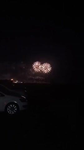 This is a vertical video taken at night. In the foreground, the left side of a silver car is visible. In the background, a dark landscape is visible with a faint horizon line. Above the landscape, fireworks are exploding in the night sky. The fireworks appear to be white and gold. The video begins with two fireworks exploding simultaneously, followed by a single larger firework, and then a scattering of smaller fireworks that fade out.