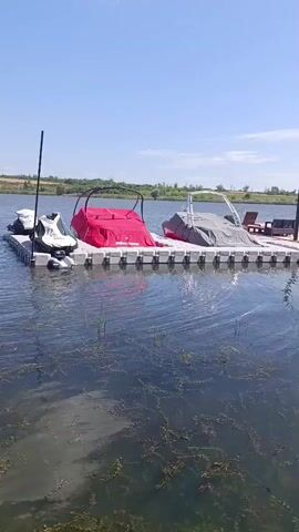 The video shows a floating dock on a body of water. The dock is made of modular plastic cubes. On the dock, there are three watercraft. On the far left, there is a white jet ski. Next to it, there is a boat covered with a red tarp. To the right of the red-covered boat, there is another boat covered with a gray tarp. A black pole is attached to the dock on the far left. In the background, there is a shoreline with trees and vegetation, and a clear blue sky with some wispy clouds. The water is calm with some areas of aquatic vegetation visible. The overall scene is one of a sunny day at a lake or calm river.