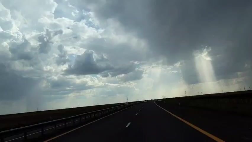 The video shows a POV shot of a road with a dark, stormy sky overhead. Sunlight beams through the clouds in several places, illuminating the otherwise dark sky. The road is asphalt and has white and yellow lines. There is a guardrail on the left side of the road, and a grassy field on either side. There are a few cars visible on the road in the distance. There are also power lines visible in the distance on the right side of the road. The overall impression is one of dramatic weather on an open road.