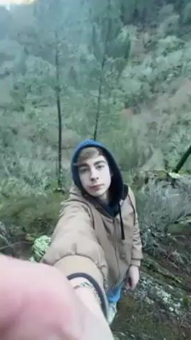 The video shows a young man taking a selfie in a mountainous, forested area. He is wearing a tan jacket, a blue hooded sweatshirt, and light-colored jeans. The man appears to be standing on a rocky outcrop. He glances at the camera then the camera moves to provide a view of the surrounding landscape. The landscape consists of steep hills covered in trees. The sky is overcast, with some muted sunlight visible through the clouds.  The overall scene suggests a natural, possibly remote location.