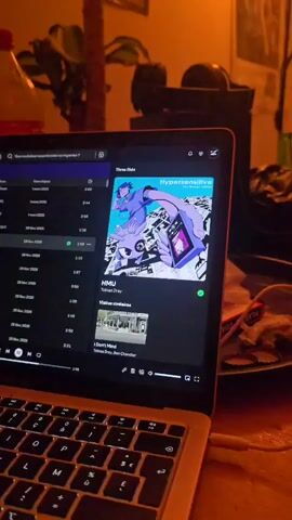 Here's a description of the video:
A laptop displays the album "Hypersensitive" by Three Kids on Spotify. The song "HMU" by Tobias Dray is playing. The laptop is on a cluttered desk with food remnants on a plate, a charging cable plugged into the side, and other objects like a coffee maker and plants in the background. The room is dimly lit, with a warm color tone.