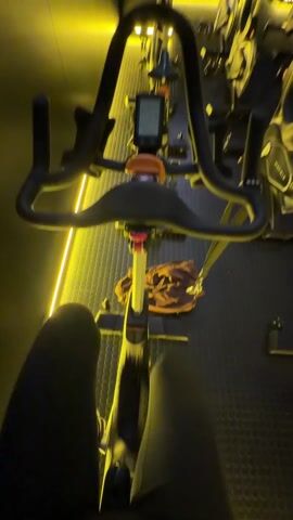 The video shows a first-person view of someone sitting on a stationary bike in a fitness studio. The person is wearing dark pants, and their legs are visible in the foreground. The bike has a digital display and an orange knob. In the background, there are multiple other stationary bikes arranged in rows. The studio has a black floor with a grid pattern and yellow lighting along the floor and on the bikes. The walls are dark, and there is a red "Emergency Exit" sign visible.