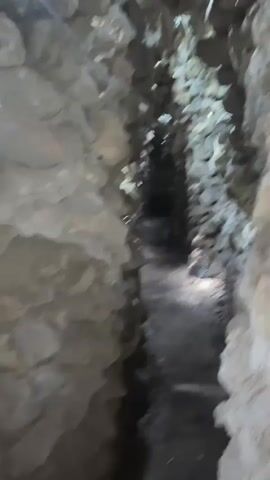 The video depicts a first-person perspective navigating a dark, narrow, and winding tunnel or passage. The walls and ceiling are constructed from rough, irregular stones, giving the impression of a cave or grotto. Dim natural light filters in from above through gaps in the stone ceiling, illuminating parts of the path ahead. The ground appears to be uneven dirt or rock.

The camera moves forward steadily, suggesting the person holding it is walking through the passage. At approximately 0:01-0:02, the person filming makes a sudden, startled sound or yelp, coinciding with an abrupt, jerky movement of the camera, as if something unexpected happened or they were surprised. No other people or specific objects are clearly visible besides the stone structure. The setting is indoors within this stone passage. The video's context indicates the location is Cam Ranh, Vietnam. No text is visible.