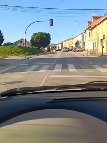 The video is shot from the dashboard of a car driving on a paved road through a residential area. The day is sunny and the sky is clear. The road has a single white line dividing it into two lanes. On either side of the road are buildings, mostly two-story houses with traditional architecture. Cars are parked along the sides of the road. In the distance, the road curves slightly to the right. A tree is visible on the left side of the road near the beginning of the street. As the car progresses, the road becomes slightly uphill. The dashboard of the car and the steering wheel are visible in the foreground. A small white piece of paper is visible on the dashboard.