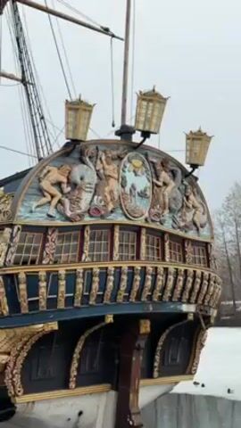 The video shows the stern of a large, ornate, historic sailing ship docked next to a body of water, partially covered in ice. The ship is dark blue and black, with elaborate gold carvings of figures and decorative elements along the stern. Three tall, ornate lanterns are mounted above the carvings.  A mast and some rigging are visible behind the stern. The water reflects the overcast sky and surrounding trees. Several ducks are swimming in the open water and standing on the ice. The video is shot with a panning motion.