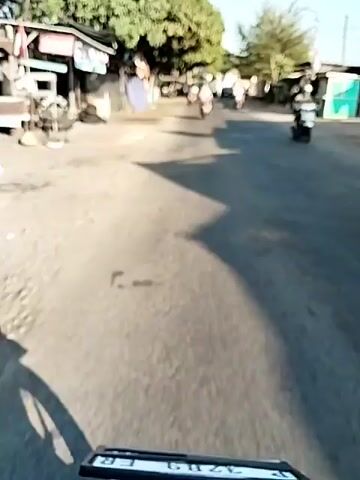 The video shows a street scene from a moving vehicle, likely a motorcycle, as indicated by the shadow of a wheel in the foreground. The road appears to be paved but somewhat worn, with patches and debris visible. On the left side of the road, there are small businesses or stalls with awnings and signage. Several trees with green foliage line the left side of the street, casting shadows on the road. On the right side, there are more structures, possibly shops or houses, and some vegetation. Several vehicles are present on the road: a few motorcycles are visible, some carrying riders, and at least one car. The sky is clear and blue, suggesting a sunny day. The overall impression is of a casual, everyday street in a town or village.