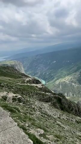 The video shows a wide shot of a lush, green canyon with a vibrant turquoise river winding through it. The camera pans from left to right, revealing the depth and vastness of the canyon. Rolling hills and mountain ranges fade into the distance under an overcast sky with dramatic clouds. A small village is nestled in the valley near the river. The foreground features a rocky, grassy slope leading down to the canyon edge, with a concrete retaining wall partially visible. The scene exudes a sense of serenity and grandeur.