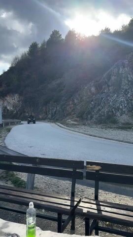 Here's a description of the video:

The video shows a black all-terrain vehicle (ATV) driving on a paved mountain road on a cloudy day. The ATV is approaching the camera, coming around a bend in the road. The sun is shining brightly behind the mountain, creating lens flare. In the foreground, there's a wooden picnic table with a bottle of water on it. The table is situated at a viewpoint, likely a rest stop. The mountain on the right has rocky outcrops and sparse vegetation.