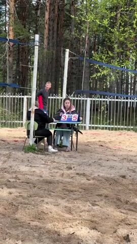 The scene features an outdoor volleyball game with the focus on the referees and a nearby volleyball court. Two women are seated at a small folding table in front of a white, metal fence, acting as referees. The woman to the right is adjusting a scoreboard, changing it from 3-5 to 3-6, while the woman to the left is taking notes. Behind them, a man in a red and black shirt stands, observing the court. The volleyball net and its poles are clearly visible, though the game itself is not the primary focus. The entire scene takes place on a sand court, surrounded by tall pine trees.