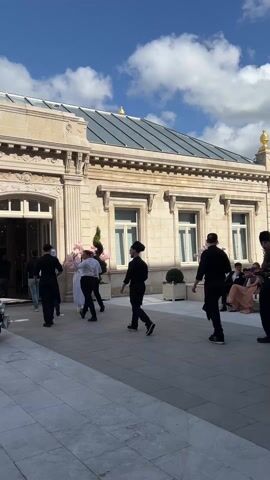 A line of people, most wearing black uniforms, walks from inside a building to an outdoor space. Some of the people in the line are holding trays and flowers. The building in the background is made of stone with a glass roof. The sky is blue with white clouds.