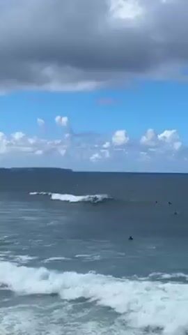 Here is a description of the video:

The video shows a surfer riding a wave in the ocean. The surfer is wearing a black wetsuit and is standing on a surfboard. The wave is breaking in front of the surfer and they are riding the wave towards the shore. There are other surfers in the water waiting for waves. The sky is blue with white and gray clouds, and there is a landmass visible in the distance. The ocean is a dark blue color, with white caps on the waves.