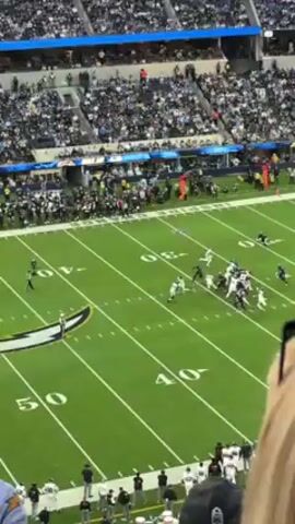 The video shows an American football game being played in a stadium, shot from a high vantage point in the stands. A group of players in black and white uniforms are actively engaged in a play on the green field, while another player in white stands nearby, appearing to survey the field. The field is marked with white yard lines and numbers. The stands are filled with spectators, many wearing team colors, creating a dense crowd. Foreground elements include the back of a man wearing a blue Chargers jersey and a woman with blonde hair. The overall atmosphere is energetic and focused on the ongoing game.