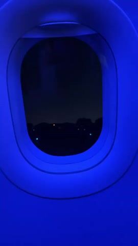 The video shows a close-up, stationary shot of an airplane window. The window frame is illuminated with a bright blue light, creating a stark contrast with the dark scene visible through the glass. Outside the window, it appears to be nighttime. A dark, silhouetted horizon line is visible, with scattered lights of various colors, including white, orange, and greenish-blue, appearing in the distance. The lights suggest some form of settlement or infrastructure on the ground below. The reflection of an overhead light fixture is visible in the upper portion of the window, and a faint reflection of a person's head can be seen in the upper left corner of the glass. The overall impression is that of someone looking out of an airplane window at night, possibly during takeoff or landing.