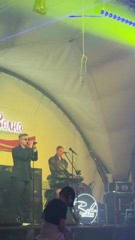This video captures a live musical performance on a stage, likely outdoors or in a large tent due to the domed ceiling structure. Two performers are prominently featured: a male singer in a black suit and sunglasses, holding a microphone and standing to the left of the stage. To his right, a female performer is seated at a keyboard, also with a microphone in front of her. Both are illuminated by stage lighting, which shifts between vibrant yellow and blue hues.

In the background, a large sign with the stylized Cyrillic word "Волна" (Volna, meaning "Wave") is visible, along with smaller text below it that appears to read "Загородный клуб" (Zagorodny klub, meaning "Country club"). A circular neon sign with the text "Roxer Cover Band" is also present on the stage, suggesting the performers are part of this group.

Several other people are in the foreground and on the stage, including children, suggesting a family-friendly event. One child in a striped shirt and another in a purple skirt are visible near the front of the stage. The overall atmosphere appears to be energetic and festive, with the dynamic lighting contributing to the mood. The specific time of day is not definitively indicated, but the use of stage lighting suggests it could be evening or nighttime. The location context provided is Tatysh, Russia.