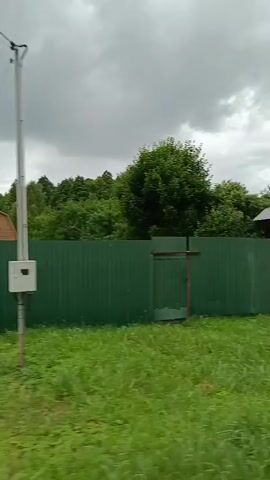 A quick shot of a rural landscape. The camera pans to the left, revealing a wooden house with ornate trim and three windows on the front. The house is surrounded by lush green foliage and flowers. Beyond the house, a dense forest can be seen. The sky is overcast, with gray clouds suggesting the possibility of rain. A green wooden fence runs across the foreground of the shot.
