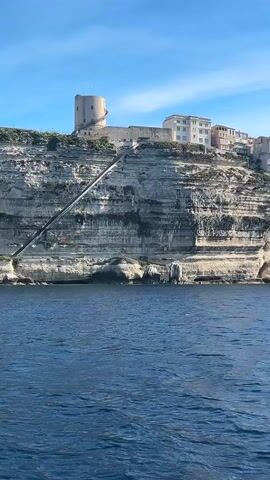 The video shows a view of a coastal town built atop white cliffs, as seen from a boat on the water. The town has a mix of buildings, including a round tower.  A long, steep staircase leads down the cliff face. The sea is a deep blue, and the sky is a clear blue with a few wispy clouds. The camera is relatively stable.