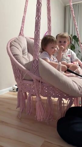 In a cozy indoor setting, two young girls are seated together in a pink macrame hanging chair. The chair is adorned with a plush pink cushion and features decorative tassels at the bottom. One girl, with light hair pulled into a ponytail, is dressed in a white t-shirt and shorts and is holding a small stuffed animal. The other girl, with short brown hair, wears a striped top. A white, fluffy pillow rests between them. The chair is gently swinging. The flooring is light wood. In the background, a desk, plant, and other toys are visible. A person, partially visible, is seated on the floor near the chair. They appear to be pushing the chair gently.