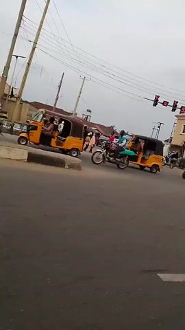 The video shows a busy intersection with several vehicles and people. A silver car is prominently in the foreground, with its passenger side visible. Multiple yellow auto-rickshaws, also known as Keke Napeps, are seen navigating the intersection. One auto-rickshaw is turning left, while others are moving straight or stopped. Several motorcycles are also present, with riders and passengers. A person in an orange vest, possibly a traffic controller or security personnel, is standing in the middle of the intersection. Pedestrians are visible on the sidewalks and crossing the street. Buildings line the background, some with shops or businesses. Overhead power lines and traffic lights are also visible. The sky is overcast, suggesting a cloudy day. The overall scene depicts a typical urban environment with active traffic flow and pedestrian activity.