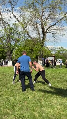 The video shows two shirtless men, wearing black pants, wrestling in a grassy park on a sunny day. A man in a blue shirt and dark pants stands between them, appearing to officiate or supervise. Several other people stand around the perimeter, watching the match. There are trees behind the wrestlers and some vehicles in the distance.